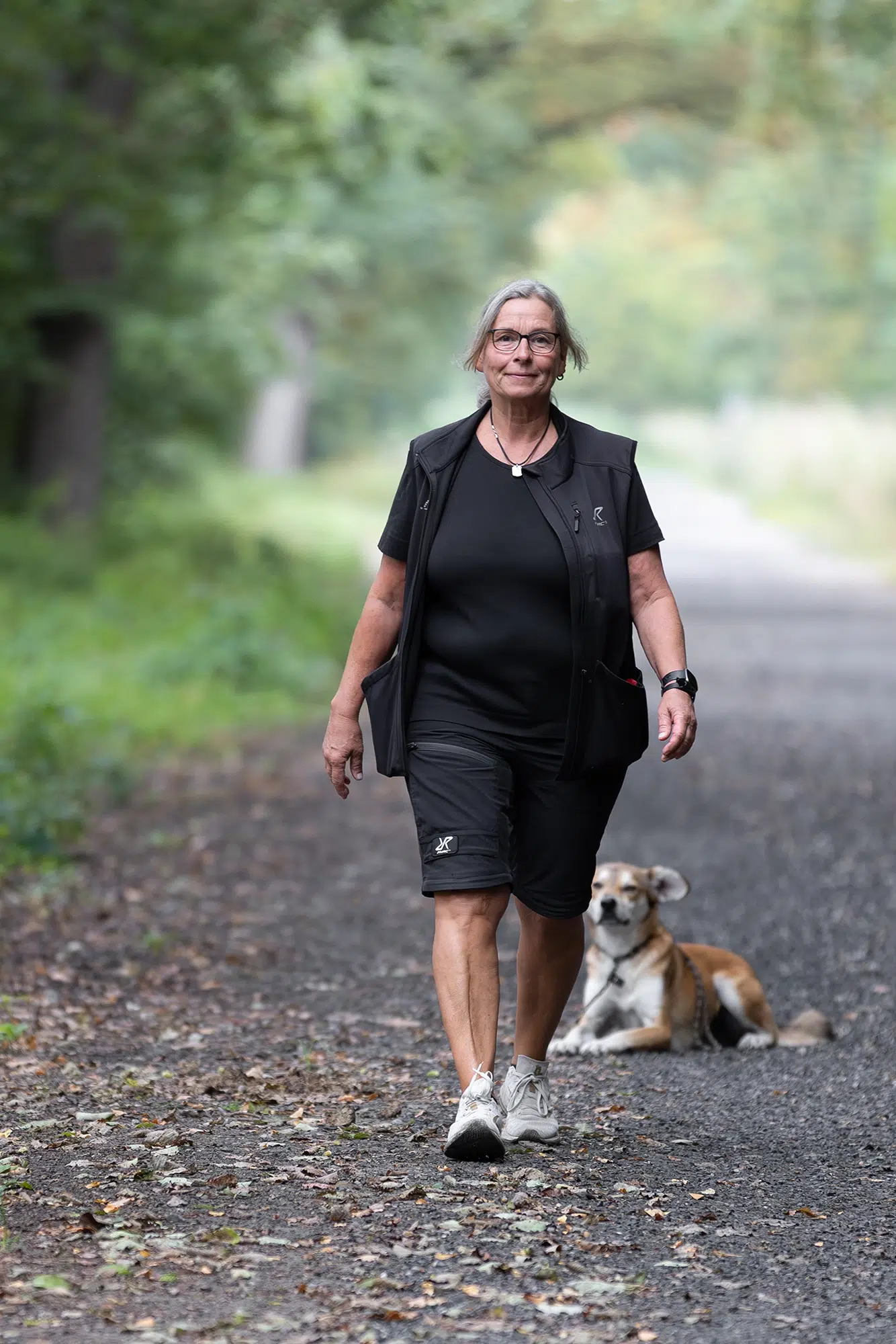 Eine Frau läuft einen Waldweg entlang, während ein Hund im Hintergrund liegt.